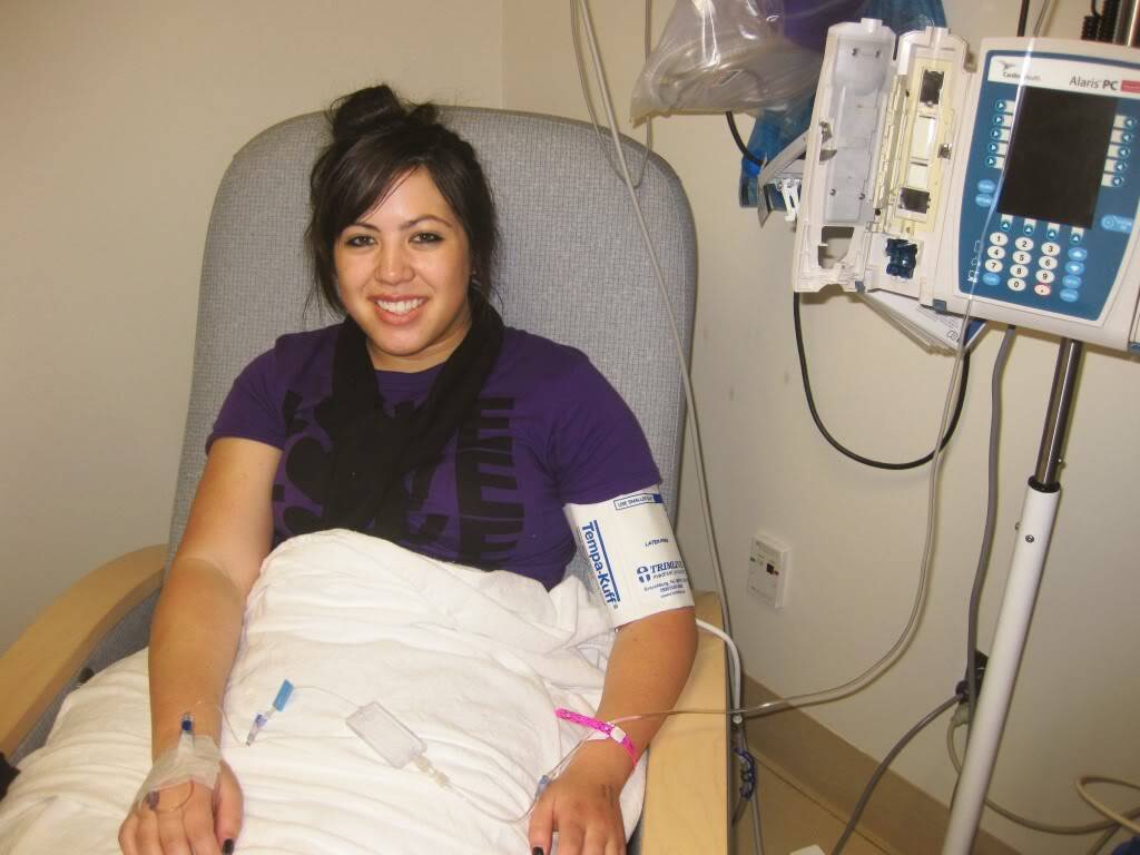 A young Amanda smiles from an infusion chair, an IV line in her hand and an infusion pump beside her.